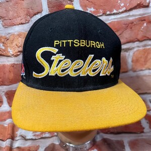 May include: Black and yellow Pittsburgh Steelers baseball cap. The cap features the team name and logo in gold lettering on the front. The brim is yellow, and the crown is black. A small team logo is on the side.