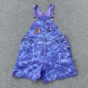 May include: Purple denim overall shorts with a unique blue splatter design. Features a front pocket with an embroidered patch and silver hardware. The straps and inner bib are a solid purple color.