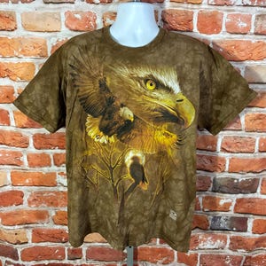 Puede incluir: Camiseta marr&oacute;n con un gr&aacute;fico detallado de un &aacute;guila, con ojos amarillos y alas extendidas. El dise&ntilde;o incluye un &aacute;guila m&aacute;s peque&ntilde;a y ramas, creando un aspecto tem&aacute;tico de la naturaleza. La camiseta se muestra en un maniqu&iacute;.