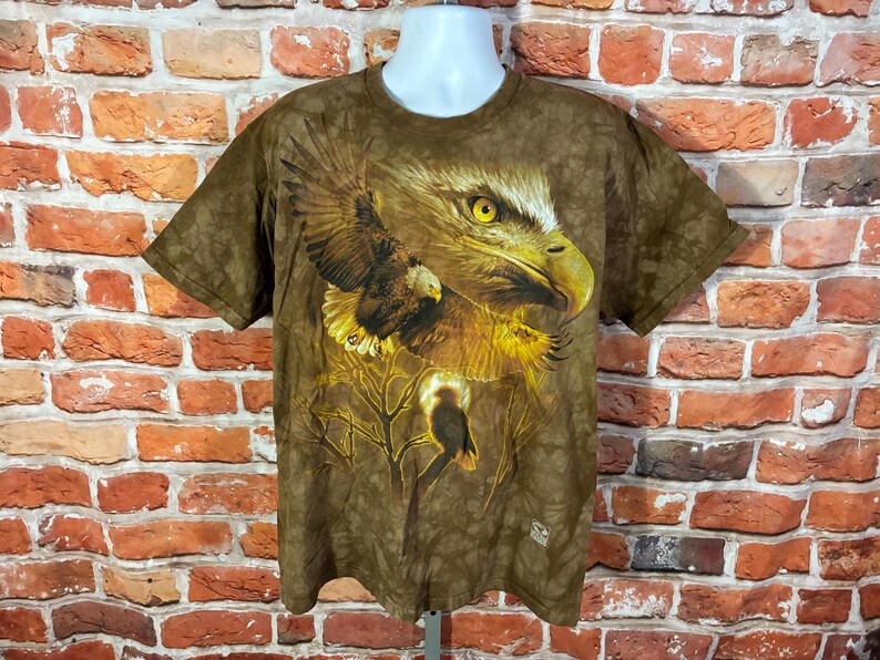 Puede incluir: Camiseta marr&oacute;n con un gr&aacute;fico detallado de un &aacute;guila, con ojos amarillos y alas extendidas. El dise&ntilde;o incluye un &aacute;guila m&aacute;s peque&ntilde;a y ramas, creando un aspecto tem&aacute;tico de la naturaleza. La camiseta se muestra en un maniqu&iacute;.