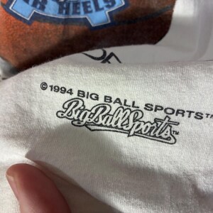 May include: White t-shirt with a basketball graphic and the text "TAR HEELS". The shirt also features the text "&copy;1994 BIG BALL SPORTS&trade;" in black. The shirt is made of a soft, comfortable material.
