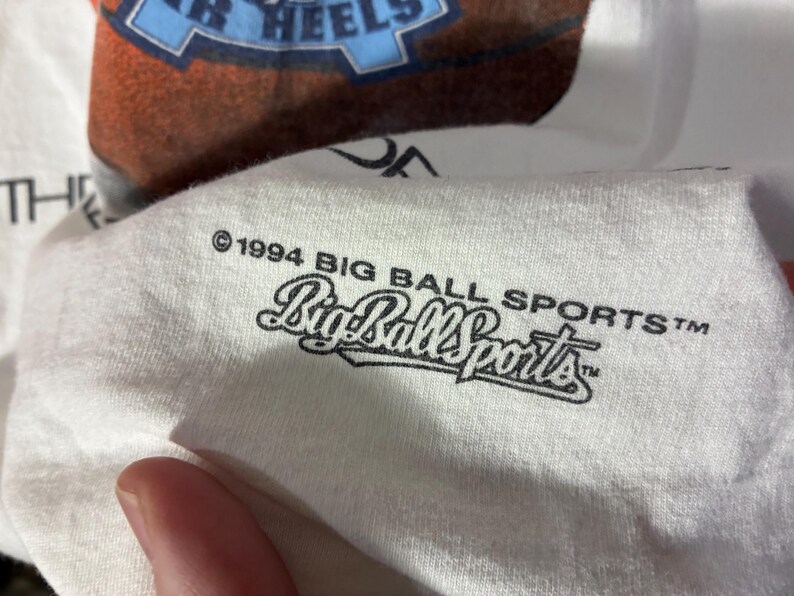 May include: White t-shirt with a basketball graphic and the text "TAR HEELS". The shirt also features the text "&copy;1994 BIG BALL SPORTS&trade;" in black. The shirt is made of a soft, comfortable material.
