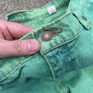 May include: Close-up of a pair of green denim jeans. The jeans have a button closure with the Levi Strauss & Co. logo. The fabric is a light green color with some darker green and blue variations. The jeans are made of a durable denim material.
