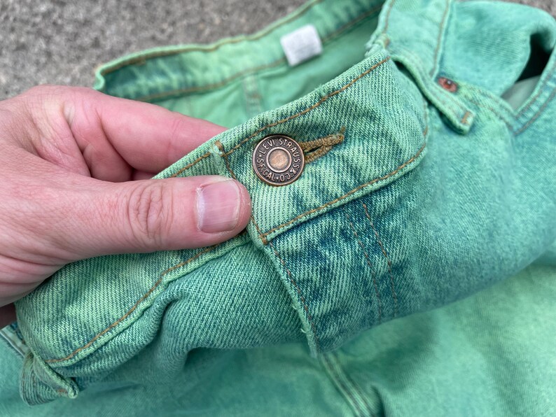 May include: Close-up of a pair of green denim jeans. The jeans have a button closure with the Levi Strauss & Co. logo. The fabric is a light green color with some darker green and blue variations. The jeans are made of a durable denim material.