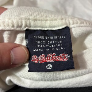 May include: Close-up of a white t-shirt collar with a navy blue tag. The tag reads "ESTABLISHED IN 1991", "100% COTTON HEAVYWEIGHT", and "MADE IN U.S.A." The brand name "Big Ball Sports" is also visible.