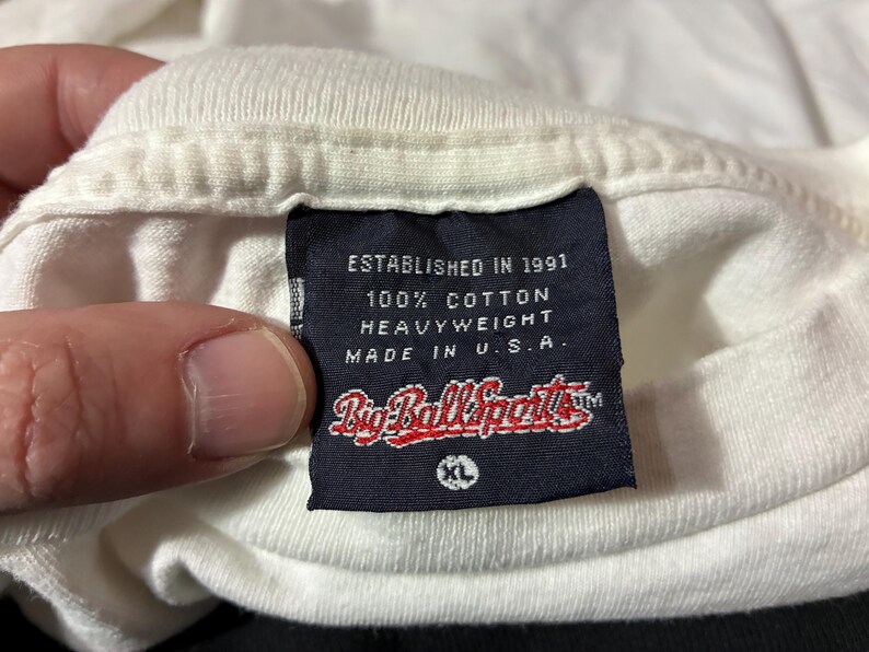 May include: Close-up of a white t-shirt collar with a navy blue tag. The tag reads "ESTABLISHED IN 1991", "100% COTTON HEAVYWEIGHT", and "MADE IN U.S.A." The brand name "Big Ball Sports" is also visible.