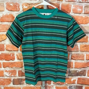 May include: A short-sleeved t-shirt with horizontal stripes in shades of green, teal, and white. The shirt is hanging on a white hanger against a brick wall background. The shirt has a crew neck and a small logo near the neckline.