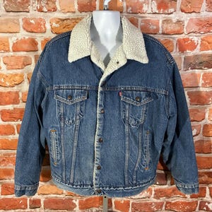 May include: A blue denim jacket featuring a cream-colored sherpa fleece collar and lining. The jacket includes two button-flap chest pockets, two side slit pockets, and a front button closure with metallic buttons and a small red brand tab.