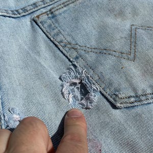Peut inclure: Gros plan sur un v&ecirc;tement en jean bleu clair avec un design us&eacute;. Le tissu pr&eacute;sente une zone d&eacute;chir&eacute;e avec des bords effiloch&eacute;s et un d&eacute;tail de poche avec des coutures en zigzag. Le v&ecirc;tement est us&eacute; et d&eacute;lav&eacute;, avec des signes d'usure visibles.