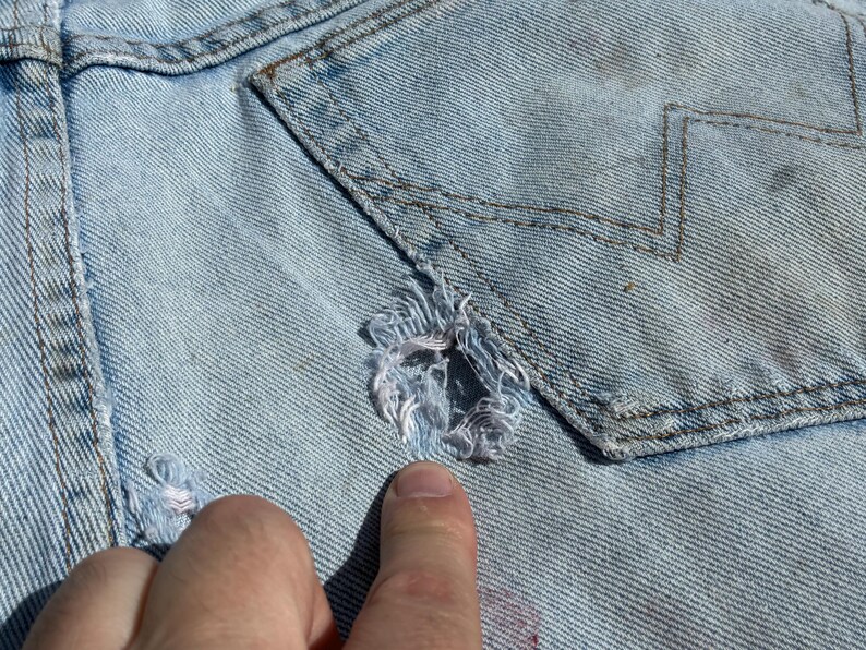 Peut inclure: Gros plan sur un v&ecirc;tement en jean bleu clair avec un design us&eacute;. Le tissu pr&eacute;sente une zone d&eacute;chir&eacute;e avec des bords effiloch&eacute;s et un d&eacute;tail de poche avec des coutures en zigzag. Le v&ecirc;tement est us&eacute; et d&eacute;lav&eacute;, avec des signes d'usure visibles.