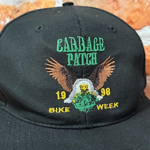 May include: Black baseball cap with embroidered text "CABBAGE PATCH" in green, an eagle design with brown wings, and the text "1998 BIKE WEEK" in yellow. The cap has a curved brim and a black button on top.