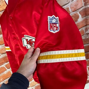 May include: Red satin jacket with white and yellow stripes on the sleeve. Features the Kansas City Chiefs logo and an NFL patch. The jacket is a vibrant red color.