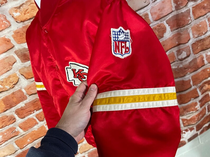 May include: Red satin jacket with white and yellow stripes on the sleeve. Features the Kansas City Chiefs logo and an NFL patch. The jacket is a vibrant red color.