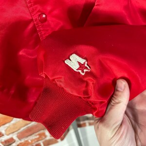 May include: A bright red satin jacket with a button closure. The jacket features a white and red star logo patch on the sleeve cuff. The cuff is a ribbed knit material.