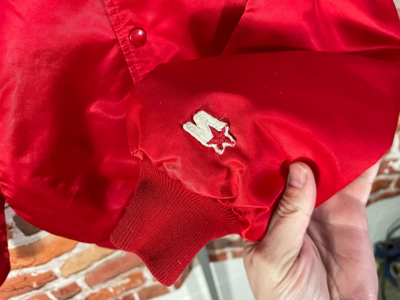 May include: A bright red satin jacket with a button closure. The jacket features a white and red star logo patch on the sleeve cuff. The cuff is a ribbed knit material.