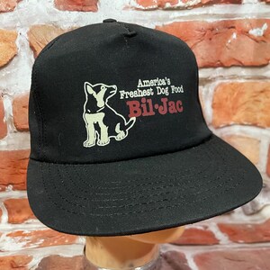 May include: Black baseball cap with a flat brim. The cap features a graphic of a dog and the text "America's Freshest Dog Food Bil-Jac" in white and red lettering. The hat is made of a durable fabric.