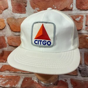 May include: A white trucker hat featuring a mesh back and a square patch on the front. The patch displays a red and orange triangle logo above the blue text CITGO, all outlined in a grey border. The hat has a solid white front panel and brim.