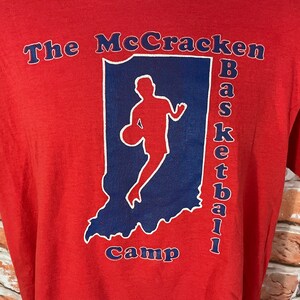 May include: Red t-shirt with a blue and white graphic of a basketball player in the shape of the state of Indiana. The text "The McCracken Basketball Camp" is printed on the shirt.