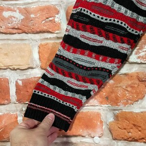 Peut inclure: Gros plan sur une manche de pull ray&eacute;e noire, rouge et grise. Le pull a une texture tricot&eacute;e et un motif g&eacute;om&eacute;trique.