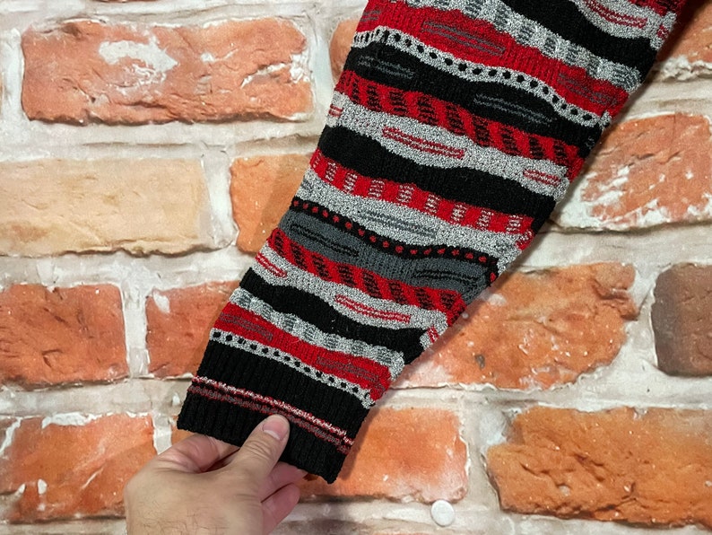Peut inclure: Gros plan sur une manche de pull ray&eacute;e noire, rouge et grise. Le pull a une texture tricot&eacute;e et un motif g&eacute;om&eacute;trique.
