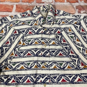 May include: A white button-down shirt with a geometric pattern in shades of blue, red, green, and brown. The pattern features triangles, leaves, and stripes.