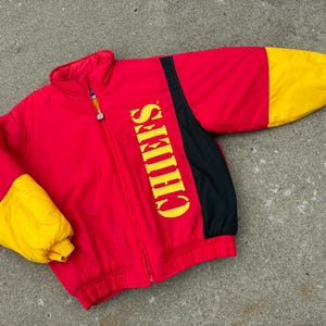 May include: A red, yellow, and black zip-up jacket featuring the word CHIEFS printed vertically in yellow on the front. The jacket has a high collar, long sleeves with yellow cuffs, and a full-length front zipper closure with a metallic pull tab.