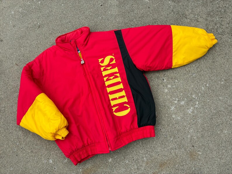 May include: A red, yellow, and black zip-up jacket featuring the word CHIEFS printed vertically in yellow on the front. The jacket has a high collar, long sleeves with yellow cuffs, and a full-length front zipper closure with a metallic pull tab.