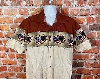 May include: Short-sleeved button-down shirt with a brown collar and upper section. The shirt features a cream-colored lower section and a patterned band with a bull, American flag, and stars design.
