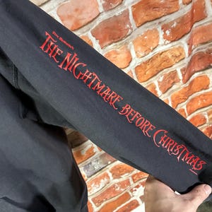 May include: Black hooded sweatshirt sleeve with the text "The Nightmare Before Christmas" in red script. The sleeve is against a brick wall background.