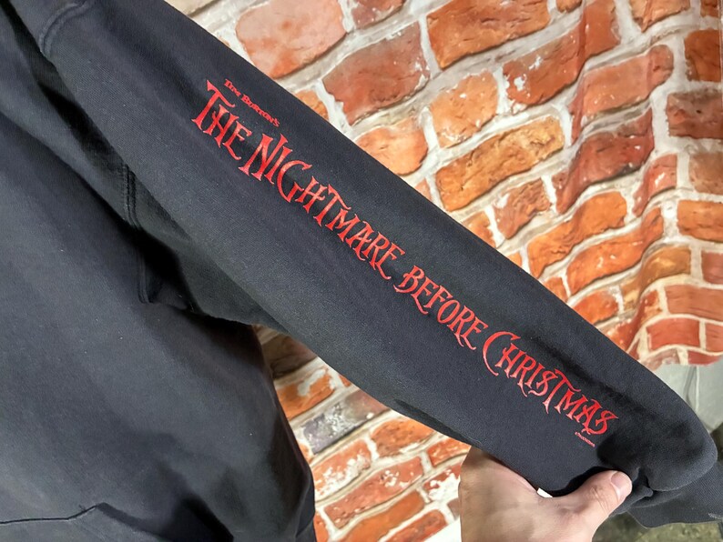 May include: Black hooded sweatshirt sleeve with the text "The Nightmare Before Christmas" in red script. The sleeve is against a brick wall background.