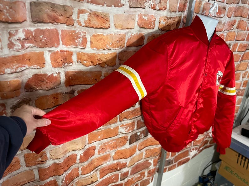May include: A red satin bomber jacket with white and yellow stripes on the sleeves. The jacket has a white patch with a logo on the chest. The jacket is displayed on a white hanger.