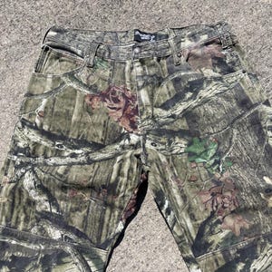 May include: Camouflage pants in shades of green, brown, and tan. The pants feature a classic five-pocket design with a button and zipper closure. The camouflage pattern is detailed with leaves and branches.