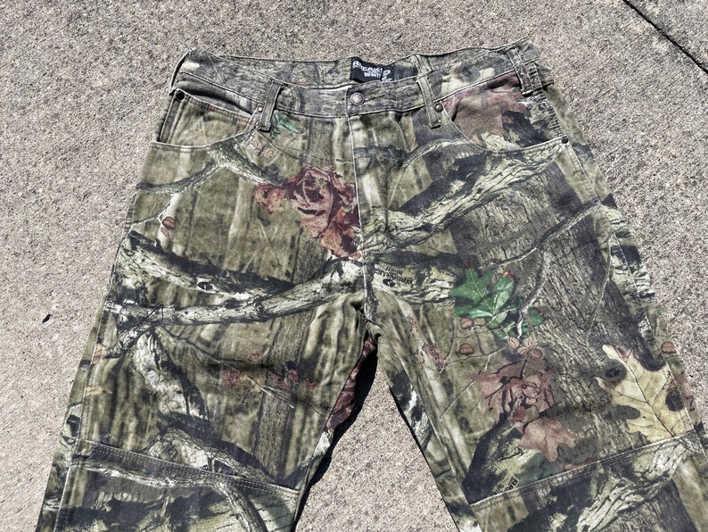 May include: Camouflage pants in shades of green, brown, and tan. The pants feature a classic five-pocket design with a button and zipper closure. The camouflage pattern is detailed with leaves and branches.