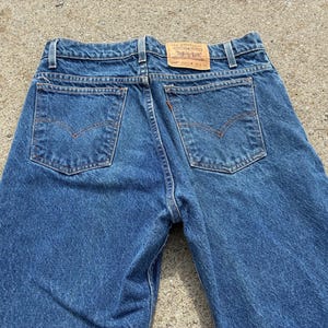 May include: Close-up of the back of blue denim jeans. The jeans have two back pockets with a classic design and a small, rectangular leather patch. The jeans are made of a medium-wash denim fabric.