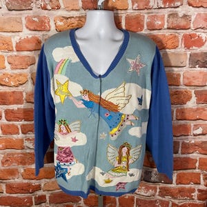 May include: A blue cardigan sweater with a whimsical design featuring embroidered fairies, stars, and clouds. The cardigan has a V-neck and a zipper closure.