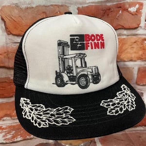 May include: A white and black trucker hat with a black mesh back. The front of the hat features a black and white illustration of a forklift with the text "Bode Finn" in red and black.