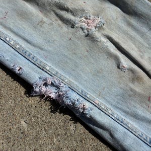 Peut inclure: Gros plan sur un jean en denim bleu clair avec des d&eacute;tails us&eacute;s. Le jean pr&eacute;sente de multiples d&eacute;chirures et des bords effiloch&eacute;s, avec des &eacute;claboussures de peinture rose et rouge visibles. Les coutures sont renforc&eacute;es par des points blancs.