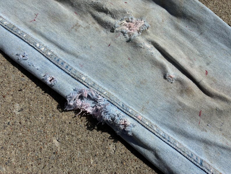 Peut inclure: Gros plan sur un jean en denim bleu clair avec des d&eacute;tails us&eacute;s. Le jean pr&eacute;sente de multiples d&eacute;chirures et des bords effiloch&eacute;s, avec des &eacute;claboussures de peinture rose et rouge visibles. Les coutures sont renforc&eacute;es par des points blancs.