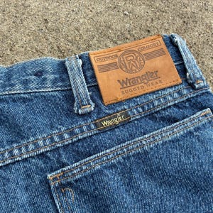 May include: Close-up of blue denim Wrangler jeans. The jeans feature a brown leather patch with the Wrangler logo and the words "Outdoor Comfort Rugged Wear." The back pocket has the Wrangler label.