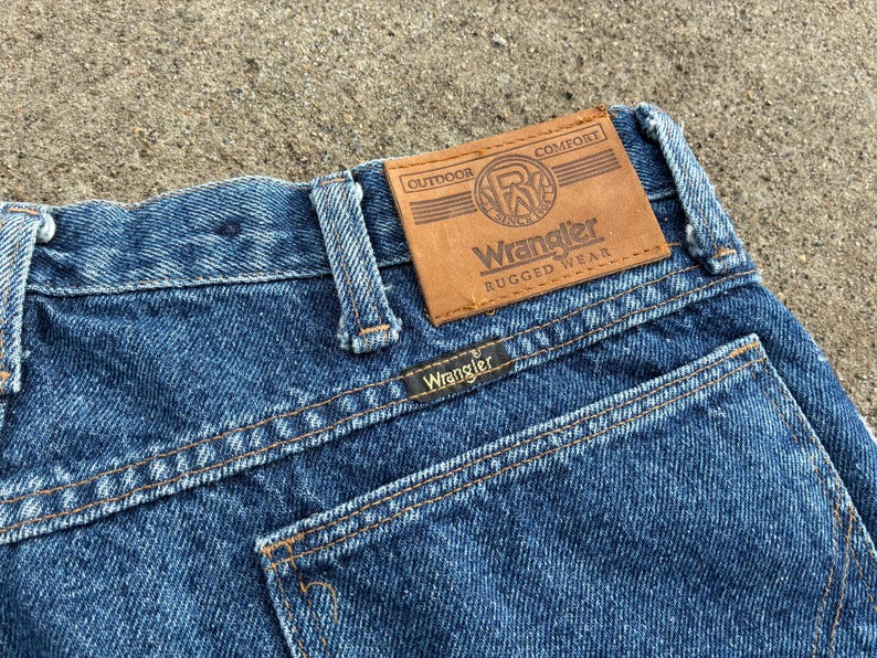 May include: Close-up of blue denim Wrangler jeans. The jeans feature a brown leather patch with the Wrangler logo and the words "Outdoor Comfort Rugged Wear." The back pocket has the Wrangler label.