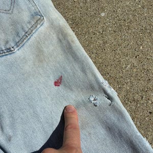 Peut inclure: Gros plan sur un jean en denim bleu clair avec une poche arri&egrave;re visible. Le jean pr&eacute;sente quelques petits trous et des taches rouges. Un doigt pointe vers le jean.