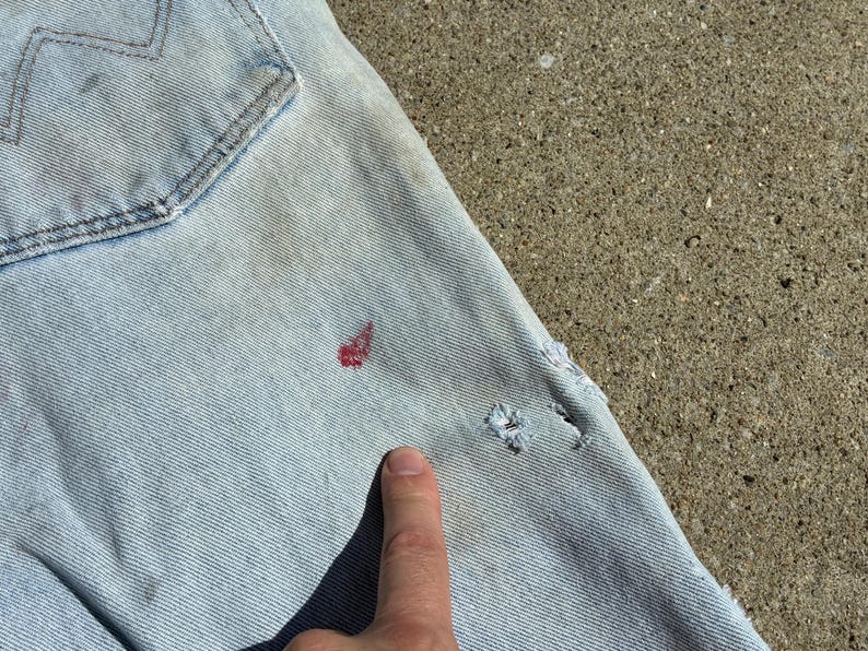 Peut inclure: Gros plan sur un jean en denim bleu clair avec une poche arri&egrave;re visible. Le jean pr&eacute;sente quelques petits trous et des taches rouges. Un doigt pointe vers le jean.