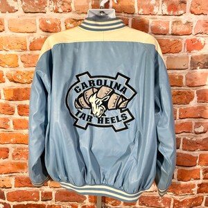 May include: Light blue and cream-colored varsity jacket with the "CAROLINA TAR HEELS" logo on the back. The jacket has a light blue body, cream-colored shoulders, and white and cream striped trim.