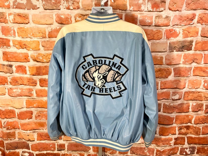 May include: Light blue and cream-colored varsity jacket with the "CAROLINA TAR HEELS" logo on the back. The jacket has a light blue body, cream-colored shoulders, and white and cream striped trim.