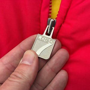 May include: A bright red jacket featuring a silver-toned metal zipper pull. The zipper pull is rectangular with a textured surface and displays the word "LOGO" above a large numeral "7" embossed in the center. The jacket has a yellow inner lining.