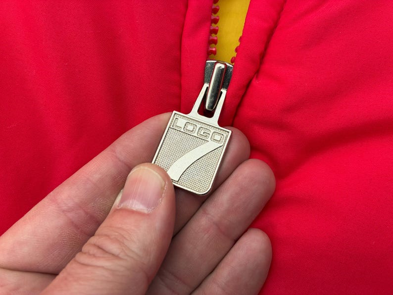 May include: A bright red jacket featuring a silver-toned metal zipper pull. The zipper pull is rectangular with a textured surface and displays the word "LOGO" above a large numeral "7" embossed in the center. The jacket has a yellow inner lining.