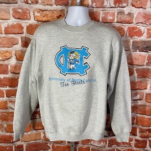Puede incluir: Sudadera gris con un logotipo azul y blanco de la Universidad de Carolina del Norte Tar Heels. El logotipo presenta una mascota de dibujos animados.