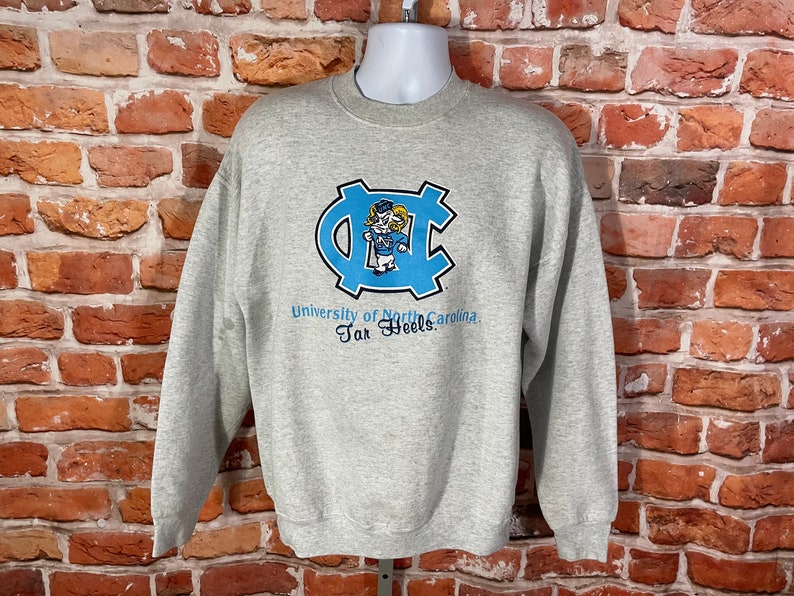 Puede incluir: Sudadera gris con un logotipo azul y blanco de la Universidad de Carolina del Norte Tar Heels. El logotipo presenta una mascota de dibujos animados.