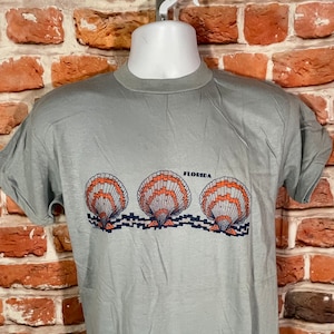 May include: A light gray t-shirt with a graphic of three orange and blue seashells with the word "Florida" printed above them.