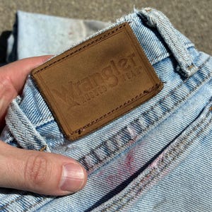 Peut inclure: Gros plan sur un jean en denim bleu clair avec une &eacute;tiquette Wrangler en cuir marron. L'&eacute;tiquette est rectangulaire avec le nom de la marque et "RUGGED WEAR" en relief. Le jean pr&eacute;sente des signes d'usure et un aspect d&eacute;lav&eacute;.